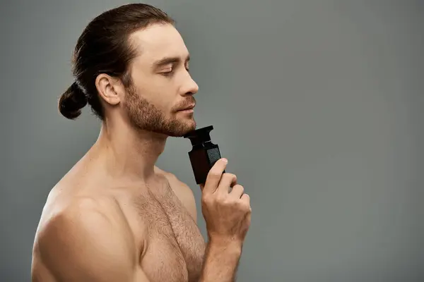 Um homem bonito e sem camisa segurando perfume em um estúdio contra um fundo cinza. — Fotografia de Stock