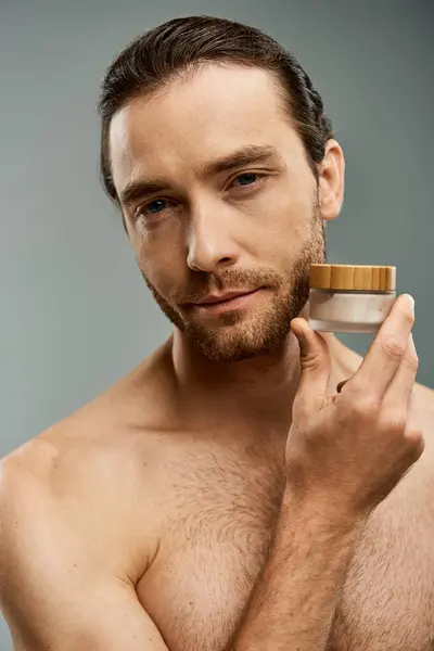 Hemdloser Mann mit Bart, der ein Glas Creme vor grauem Hintergrund im Studio hält. — Stockfoto
