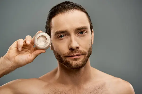 A shirtless, bearded man holding a jar of cream in his hand against a grey studio backdrop. — Stock Photo