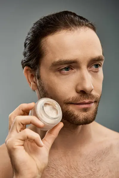 Un hombre sin camisa con barba sosteniendo delicadamente un frasco de crema y aplicándolo en la cara contra un fondo gris de estudio. - foto de stock