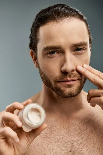 Handsome shirtless man with a beard holding a jar of cream against a grey backdrop. — Stock Photo