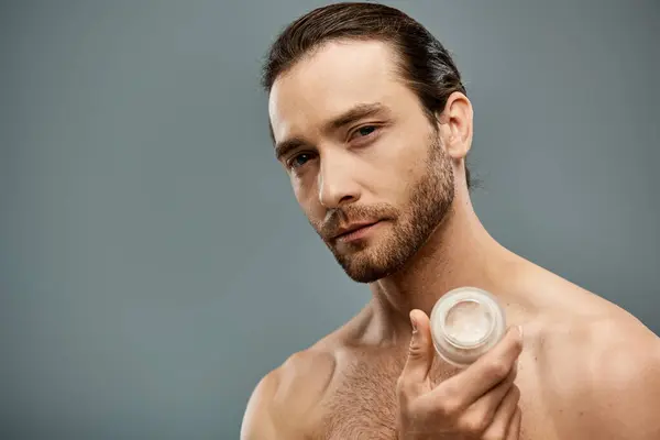 Un hombre guapo, sin camisa con una barba sosteniendo crema en la mano contra un fondo gris del estudio. - foto de stock