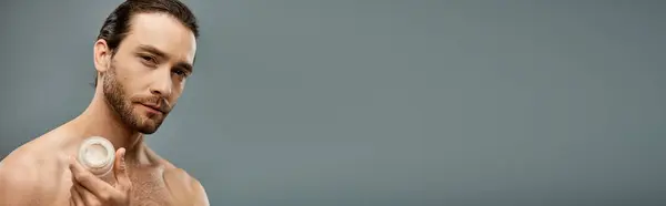 Handsome shirtless man with a beard holding a jar of cream against a grey background — Stock Photo