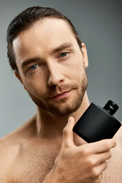A handsome shirtless man with a beard holds perfume bottle against a grey background in a studio setting. — Stock Photo