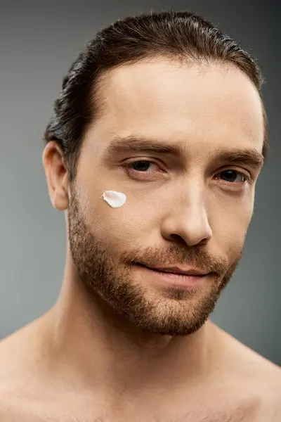 Un hombre guapo y sin camisa con barba se pone una crema en la cara en un estudio. - foto de stock
