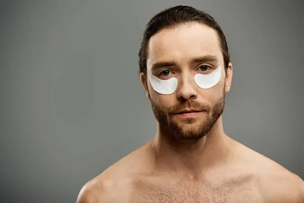 Un bel homme torse nu avec une barbe porte avec confiance des bandeaux dans une pose frappante sur un fond de studio gris. — Photo de stock