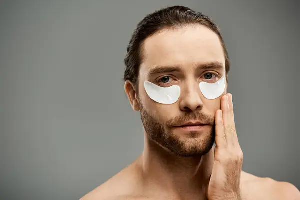A handsome and shirtless man with a beard wearing eye patches on his face in a studio against a grey background. — Stock Photo