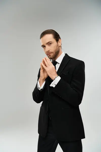 Un homme d'affaires barbu en costume et cravate se tient les mains jointes dans une pose réfléchie sur un fond gris. — Photo de stock