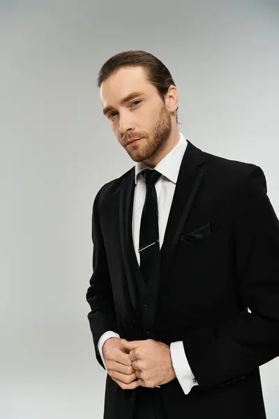 Un homme d'affaires charismatique et barbu en costume pointu et cravate frappe une pose confiante sur un fond de studio gris. — Photo de stock