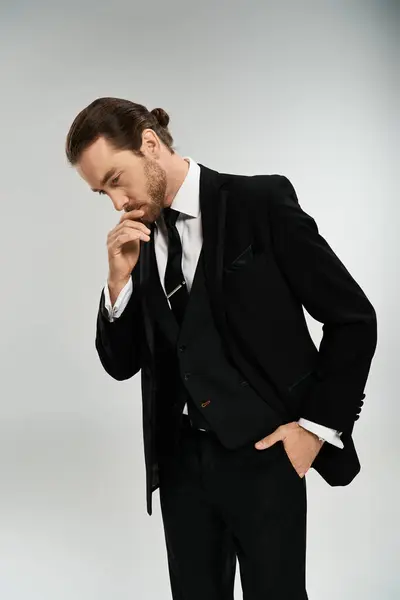 Un hombre de negocios barbudo con un traje elegante toma una postura confiada contra un fondo gris del estudio. - foto de stock