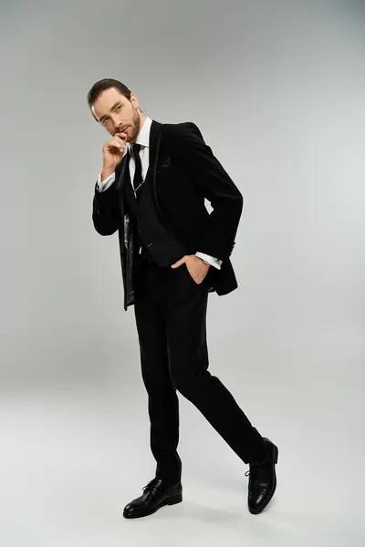 Un hombre de negocios guapo y barbudo posa elegantemente en un esmoquin contra un fondo gris de estudio. - foto de stock