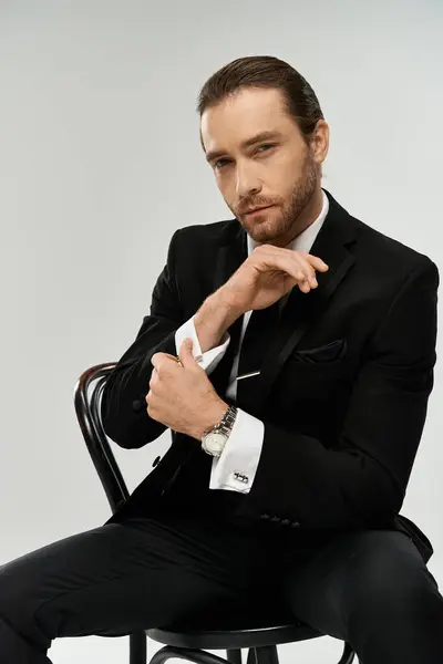 Ein bärtiger Geschäftsmann sitzt auf einem Stuhl und strahlt Charisma und Selbstvertrauen in seinem maßgeschneiderten Anzug vor grauem Studiohintergrund aus.. — Stockfoto