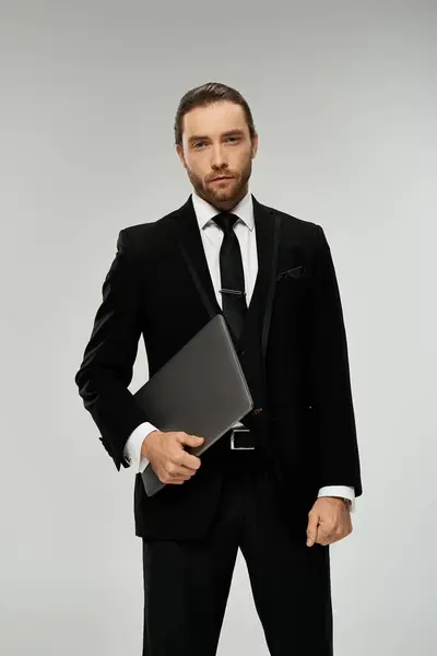 A handsome, bearded businessman in a suit confidently holds a laptop in a studio set against a grey background. — Stock Photo