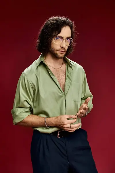 Un homme élégant et beau en lunettes, portant une chemise en soie verte, frappant une pose sur un fond rouge vif. — Photo de stock