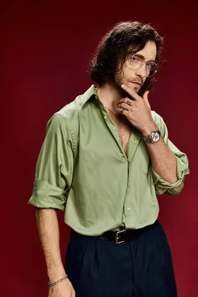 Schöner Mann mit Brille posiert in grünem Seidenhemd und schwarzer Hose vor leuchtend rotem Hintergrund. — Stockfoto