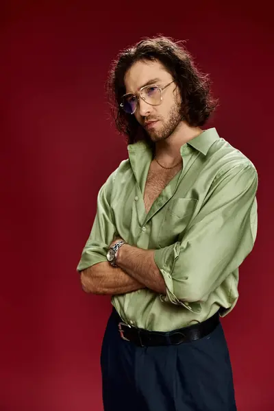 Ein gutaussehender Mann mit verschränkten Armen, Brille und grünem Seidenhemd, der selbstbewusst auf rotem Grund posiert. — Stockfoto