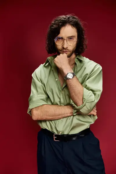 A handsome man in glasses and a green silk shirt showcases a luxurious watch on his left hand against a bold red background. — Stock Photo