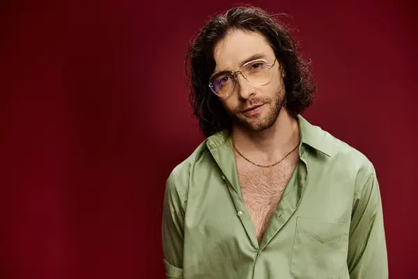 A handsome man wearing glasses and a green silk shirt poses against a vibrant red background. — Stock Photo