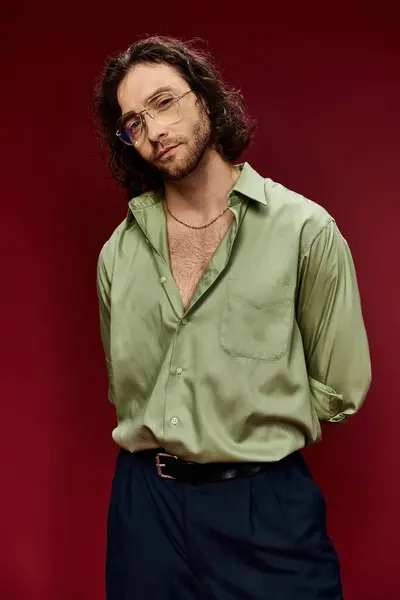 Un homme charismatique aux cheveux longs et aux lunettes portant une chemise de soie verte pose gracieusement sur un fond rouge vif. — Photo de stock
