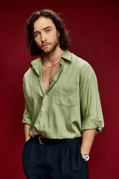 Un homme captivant aux cheveux longs pose dans une chemise de soie verte sur fond rouge frappant. — Photo de stock