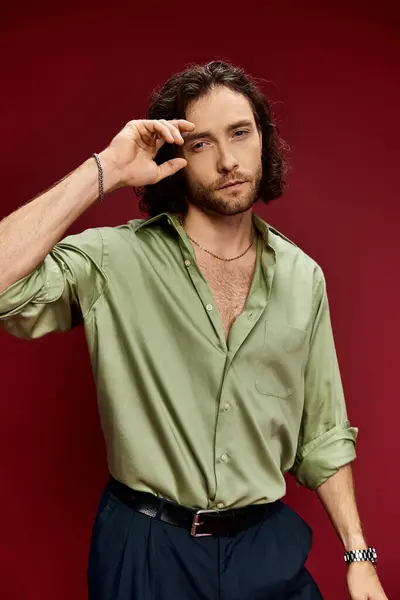 Красивий чоловік з довгим волоссям і зеленою шовковою сорочкою впевнено позує на яскравий червоний фон. — стокове фото