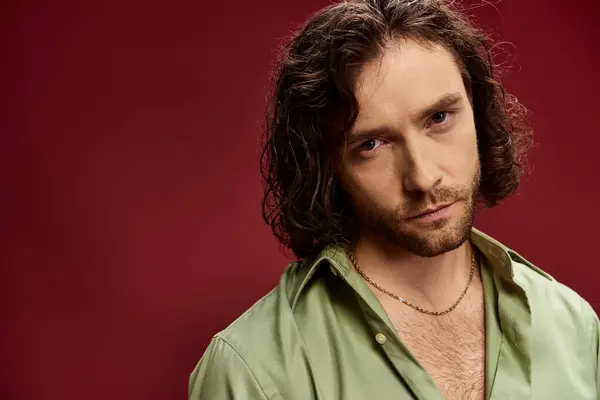 A stylish man with long hair and glasses strikes a pose in a vibrant green silk shirt on a bold red backdrop. — Stock Photo