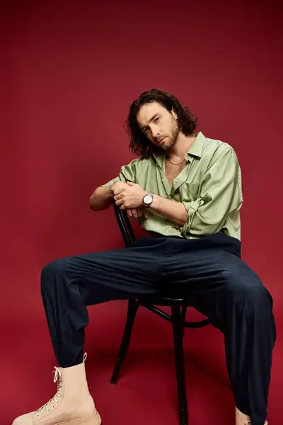 A handsome man in glasses and a green silk shirt sits with legs crossed on a chair against a vibrant red backdrop. — Stock Photo