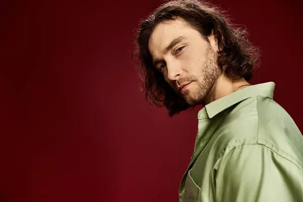 Handsome man with long hair and glasses striking a pose in a green shirt on a vibrant red backdrop. — Stock Photo