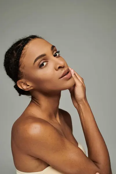 Jeune femme afro-américaine dans un haut sans bretelles posant gracieusement avec sa main sur sa joue sur un fond gris. — Photo de stock