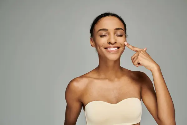 Una giovane donna afroamericana trasuda fiducia, sorridendo in un reggiseno senza spalline mentre si prende cura della sua pelle su uno sfondo grigio. — Foto stock