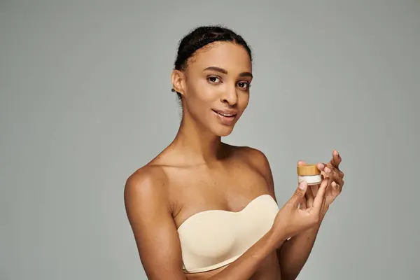 Young African American woman in strapless bra holding cream on grey background. — Stock Photo