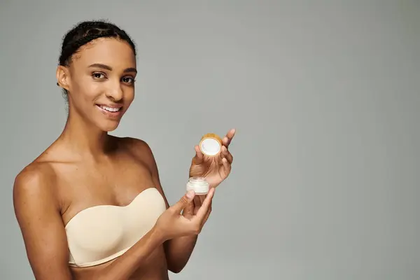 Young African American woman in a bra taking care of her skin, holding a container of cream on a grey background. — Stock Photo