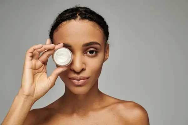 Junge Afroamerikanerin im schulterfreien Top hält Cremedose vor Gesicht, Hautpflegekonzept, grauer Hintergrund. — Stockfoto