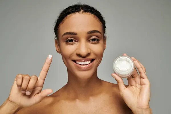 Young African American woman in strapless top holding jar of cream in front of face, skin care routine on grey background. — Stock Photo