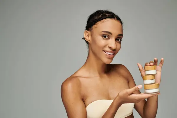 A young African American woman holding cream jars on a grey background. — Stock Photo