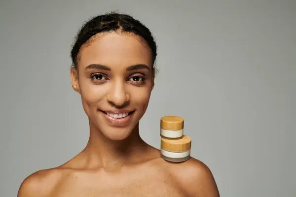African American woman in strapless top smiles, beauty jars with cream on grey background. — Stock Photo