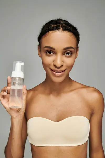 Giovane donna afroamericana in reggiseno, tenendo detergente per il viso, mostrando routine di cura della pelle su sfondo grigio. — Foto stock