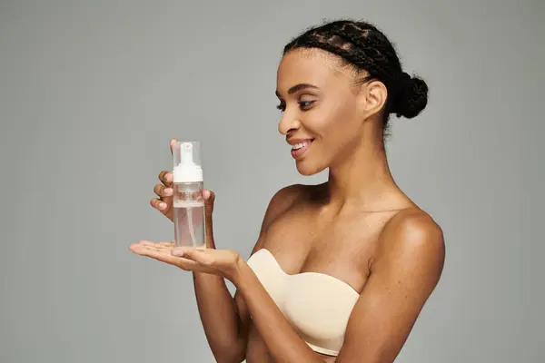 Молода афроамериканка в бюстгальтері тримає пляшку засобу для чищення обличчя, піклуючись про її шкіру. — стокове фото