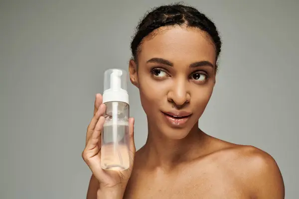 A young African American woman in a strapless top holds a bottle of liquid in front of her face on a grey background. — Stock Photo