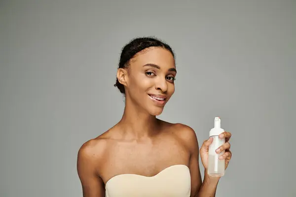 Eine junge Afroamerikanerin mit makelloser Haut hält eine Flasche Gesichtsreiniger anmutig und ausbalanciert vor grauem Hintergrund. — Stockfoto