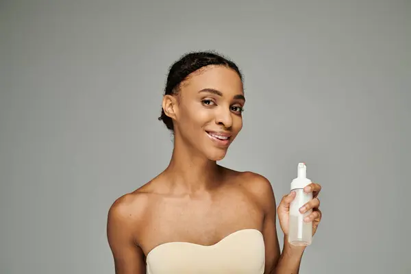 A young African American woman with flawless skin holds a bottle of facial cleanser in a graceful and poised manner on a grey background. — Stock Photo