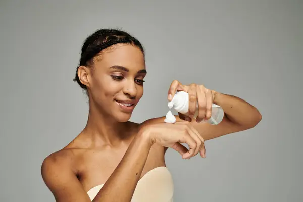 A young African American woman, wearing a strapless top, holding facial cleanser against a grey backdrop. — Stock Photo