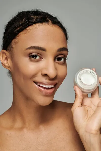 Mulher afro-americana em top strapless detém frasco de creme, cuidando da pele no fundo cinza. — Fotografia de Stock