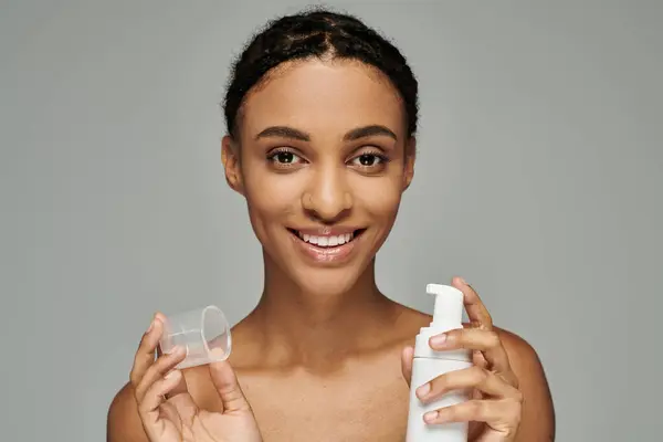 Junge Afroamerikanerin in trägerlosem Top hält Flaschen Reinigungsmittel in der Hand und pflegt ihre Haut auf grauem Hintergrund. — Stockfoto