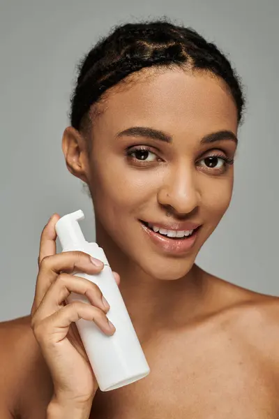 Young African American beauty in strapless top delicately holds a bottle of cleanser on a grey background. — Stock Photo