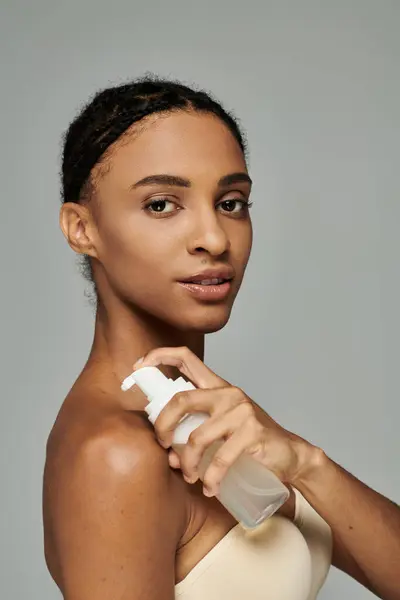 Junge Afroamerikanerin mit einer Flasche Reinigungsmittel, um die Haut mit Feuchtigkeit zu versorgen, trägt schulterfreies Oberteil auf grauem Hintergrund. — Stockfoto