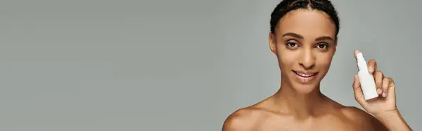 Young African American woman in strapless top gently holding a white skincare product in her right hand against a grey background. — Stock Photo