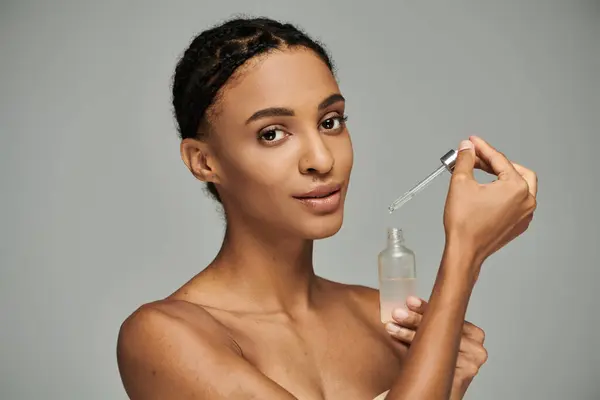 African American woman in a strapless top gracefully holds a bottle of serum against a grey background. — Stock Photo