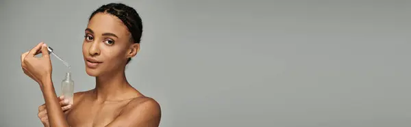 Молодая афро-американка в рубашке без бретелек держит в руках бутылку сыворотки, обнимая на сером фоне свою рутину с кожей.. — стоковое фото