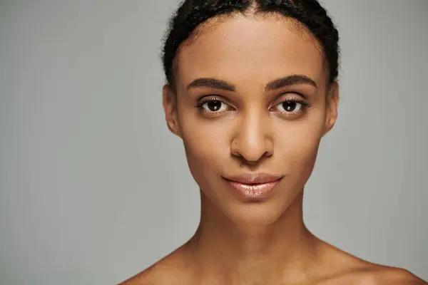 Una bella e giovane donna afroamericana in un top senza spalline che si prende cura meticolosamente della sua pelle su uno sfondo grigio. — Foto stock